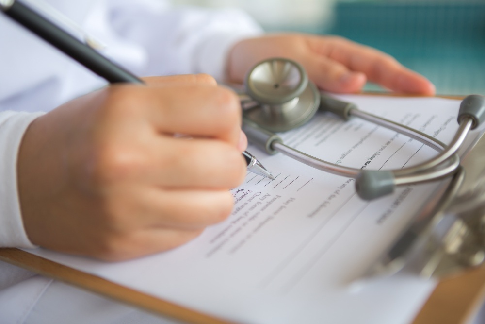Profissional de saúde assinando documento médico com estetoscópio ao lado.